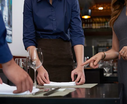 Een restaurantsetting met drie mensen die een tafel dekken. Iemand in een blauwe shirt houdt een servet vast, terwijl een ander in grijs wijst. Elegante, gefocuste sfeer.