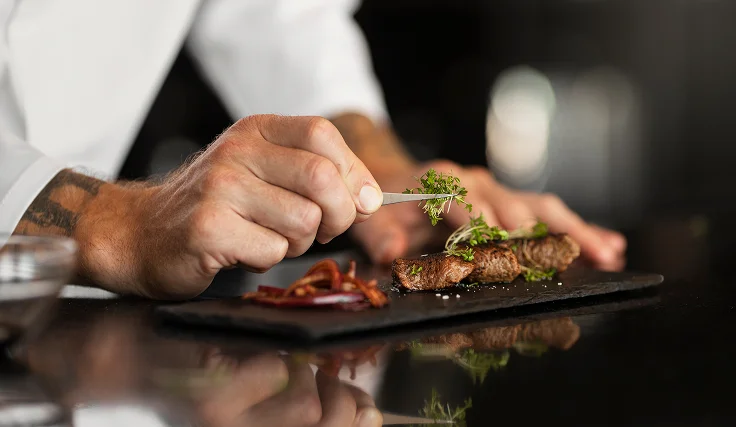 Een chef-kok garneert een steak met verse kruiden op een zwart leisteen bord. De focus ligt op de precieze handbewegingen, wat een gevoel van kunstzinnigheid en elegantie oproept.