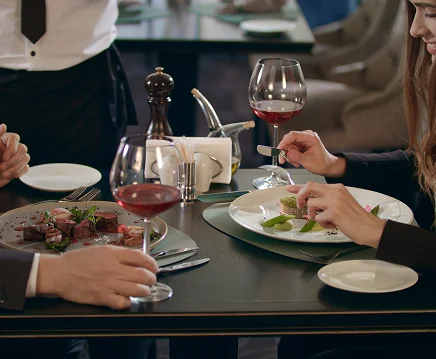 Een stel dineert in een restaurant, zittend aan een tafel met elegante gerechten en rode wijn. Beiden zijn betrokken in een gesprek, wat een gezellige, intieme sfeer uitstraalt.